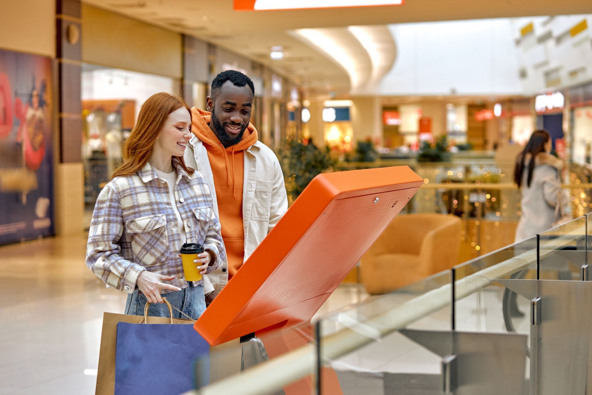 Two shoppers reading an orange mall directory kiosk in a bright indoor shopping center.