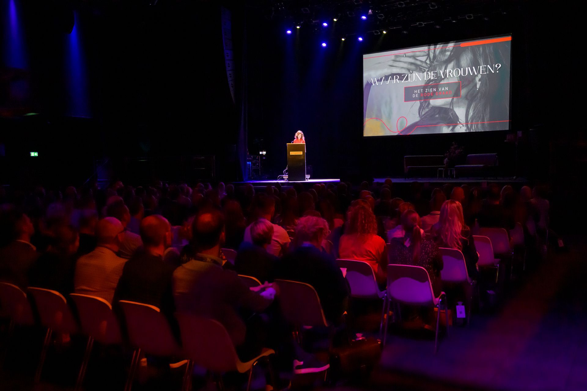 Het publiek zit in een schemerig verlicht theater en kijkt naar een spreker op het podium naast een groot scherm.