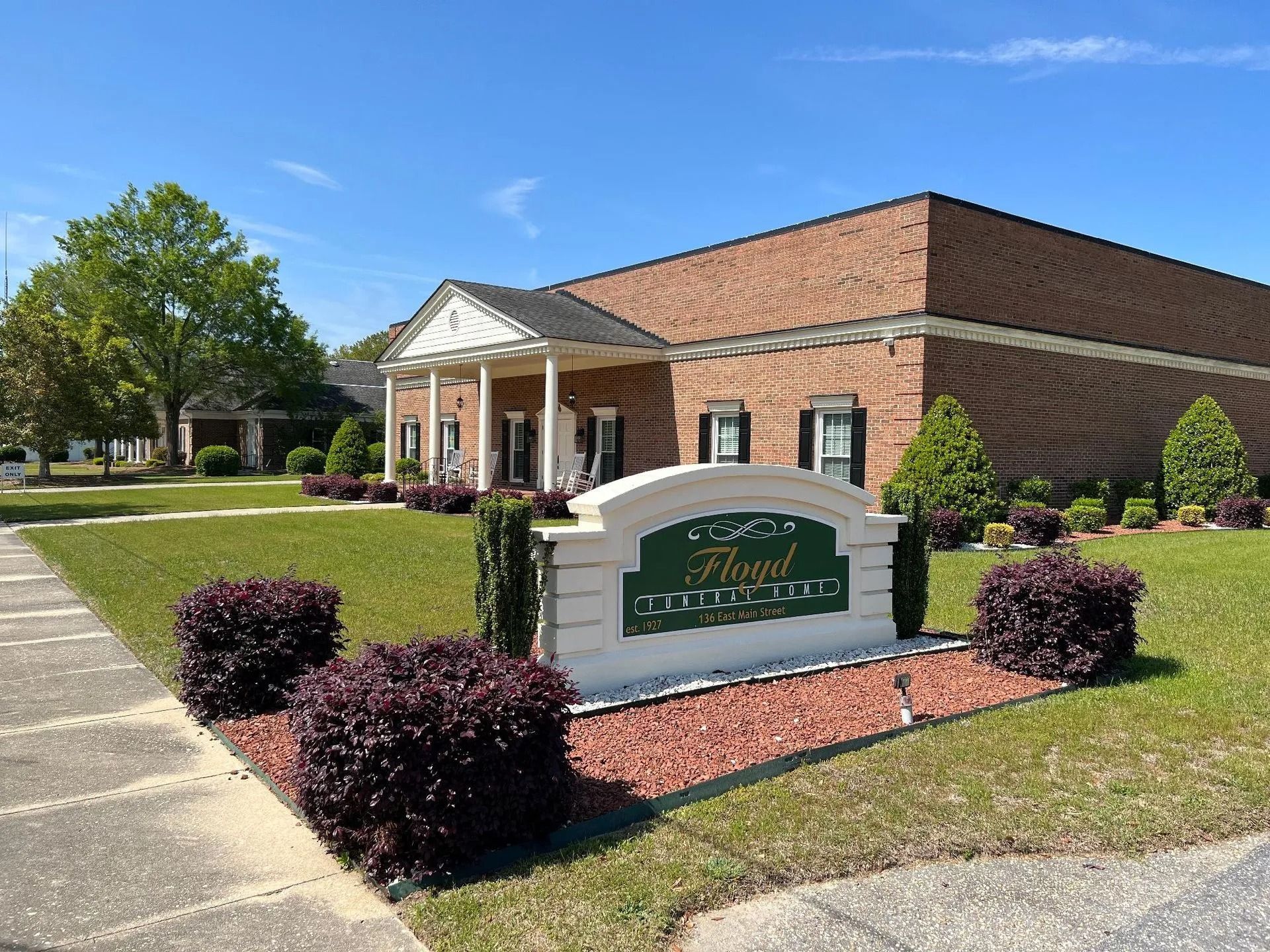 Sign in front of a brick building with a colonnaded entrance; landscaping.