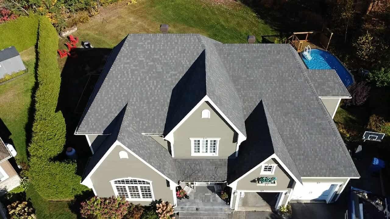 Vue aérienne d'une maison au toit gris et aux boiseries blanches, entourée de verdure et d'une couverture de piscine.