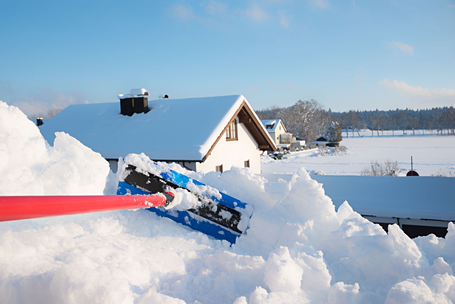 Par une journée d'hiver ensoleillée, une personne déneige un toit à l'aide d'une pelle bleue.