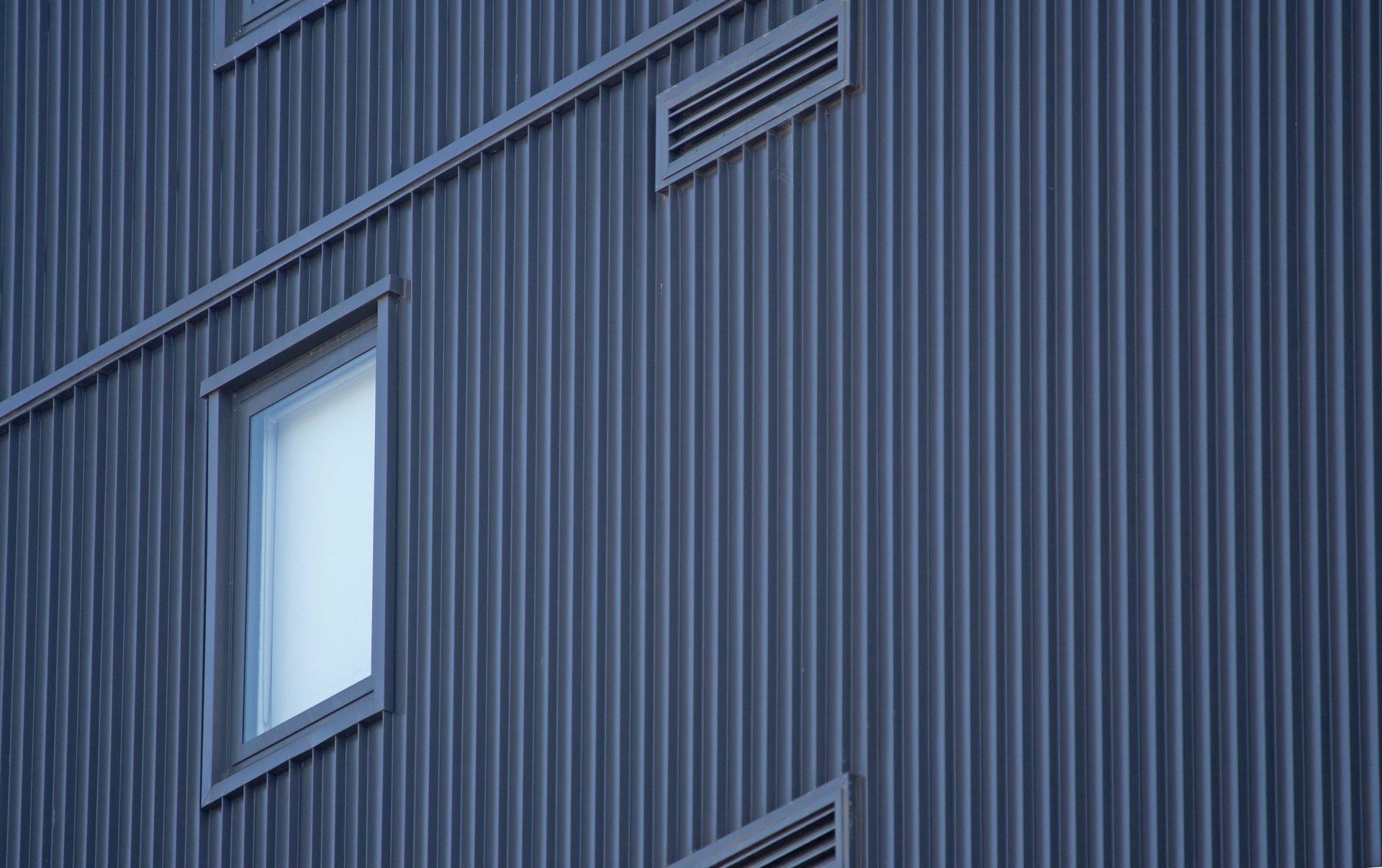 Extérieur du bâtiment en tôle ondulée gris foncé, avec une fenêtre et des grilles de ventilation.