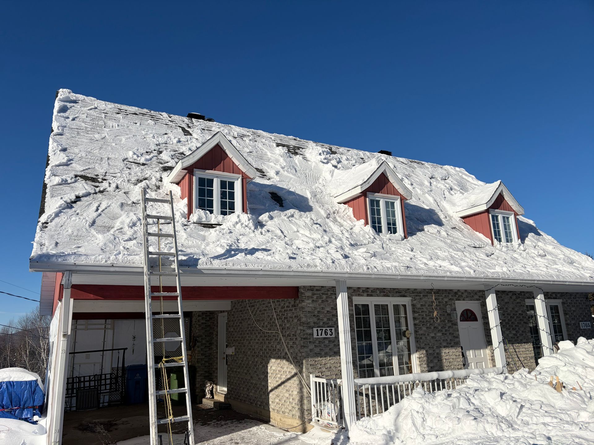 Maison enneigée aux boiseries rouges, avec trois lucarnes et une échelle appuyée contre le toit par une journée ensoleillée.