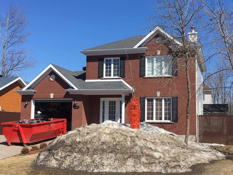 Maison en briques de deux étages avec un gros tas de neige sale devant et une benne à ordures rouge dans la cour.