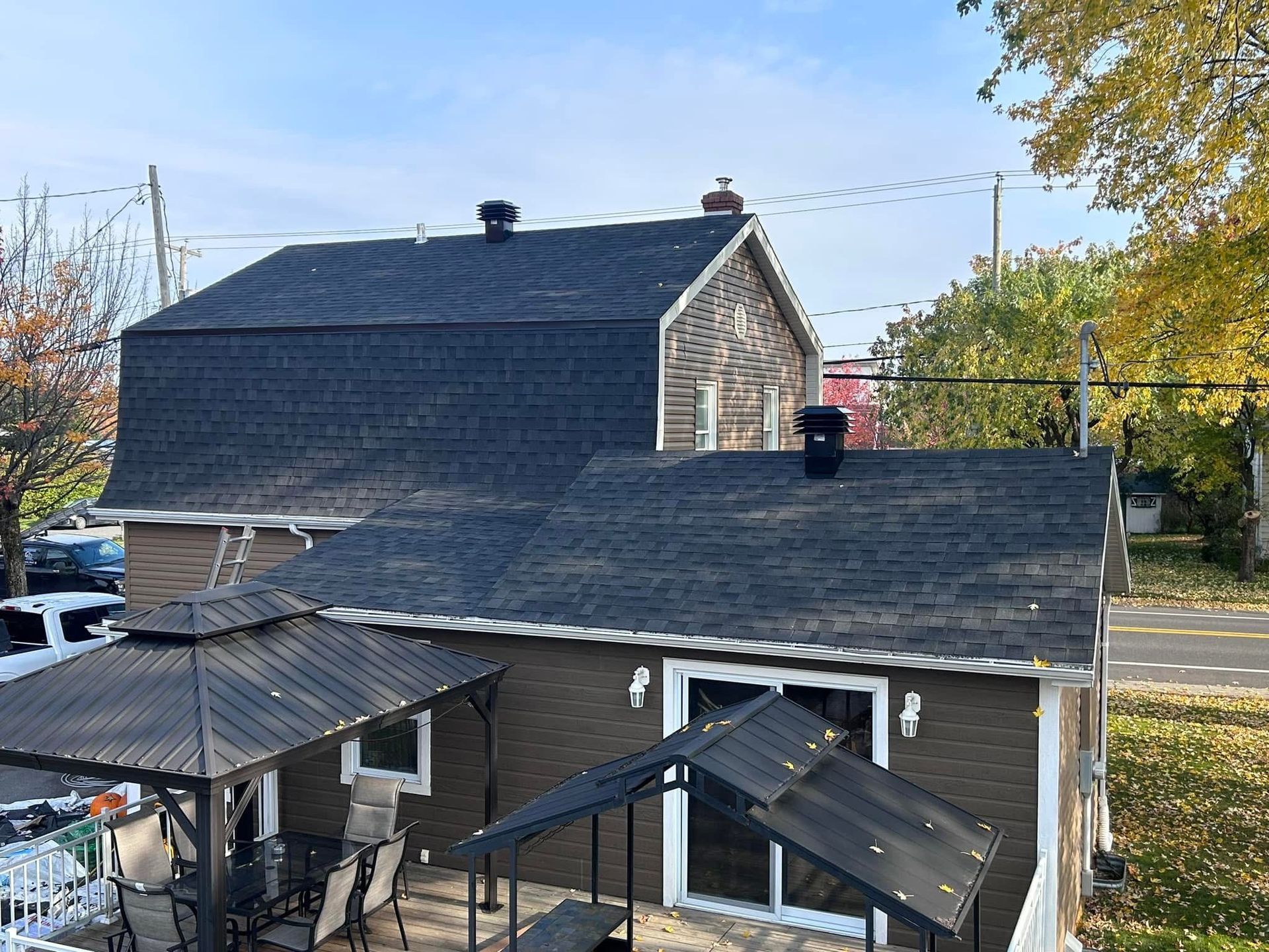 Maison brune au toit noir, terrasse avec gazebo. Arbres d'automne et ciel bleu en arrière-plan.