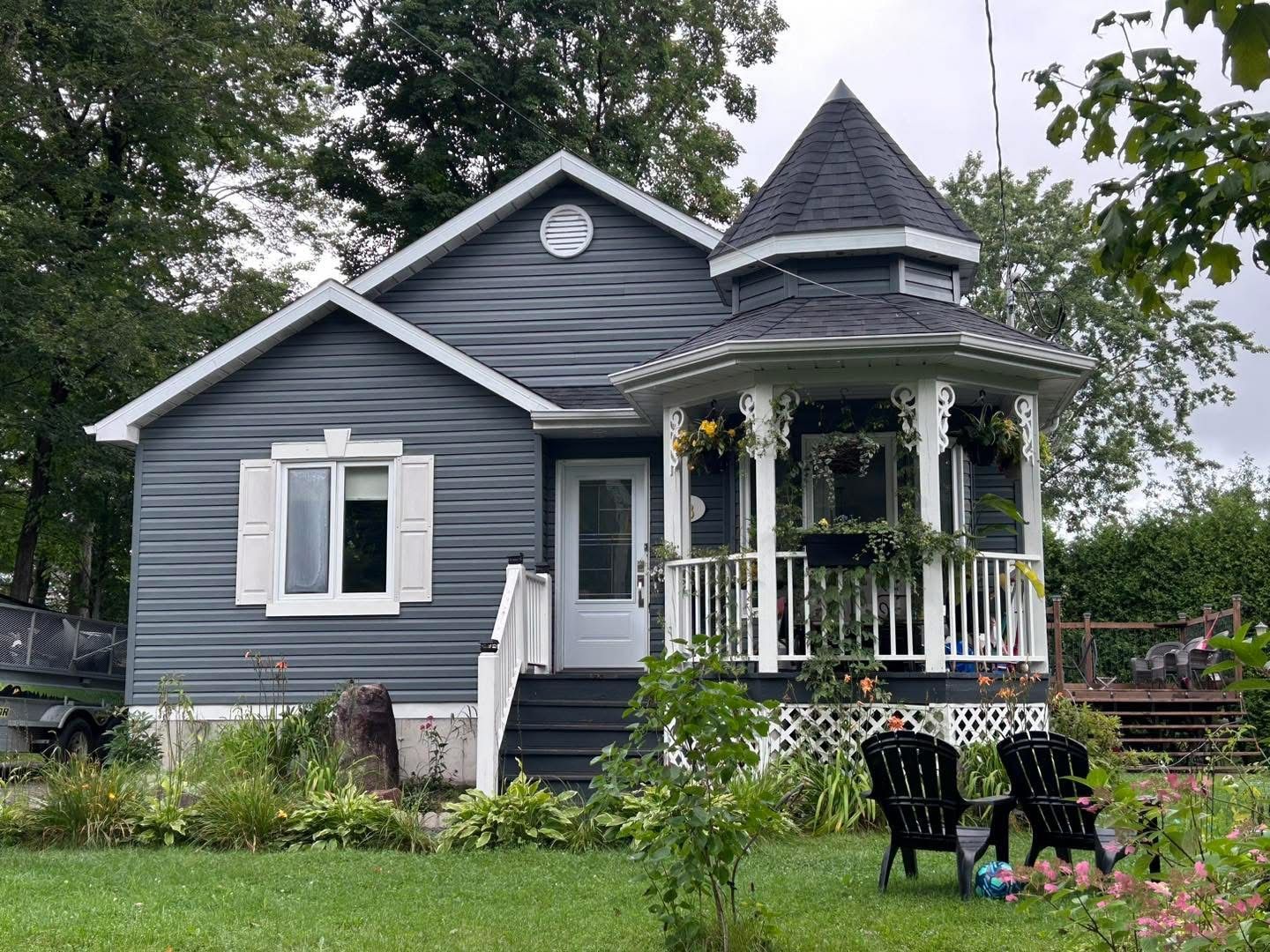 Maison bleu-gris avec une véranda de style gazebo. Volets et boiseries blanches, chaises noires sur la pelouse, feuillage vert.