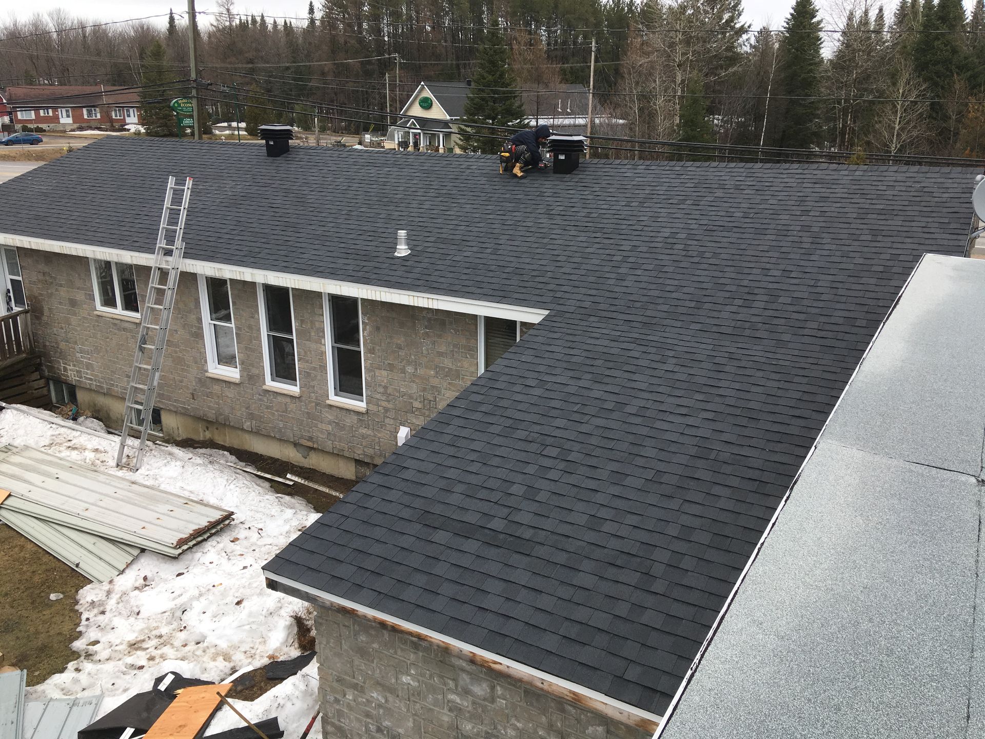 Vue d'une maison dont le toit est fraîchement recouvert de bardeaux noirs. Une échelle est appuyée contre le mur et des ouvriers travaillent sur le toit.