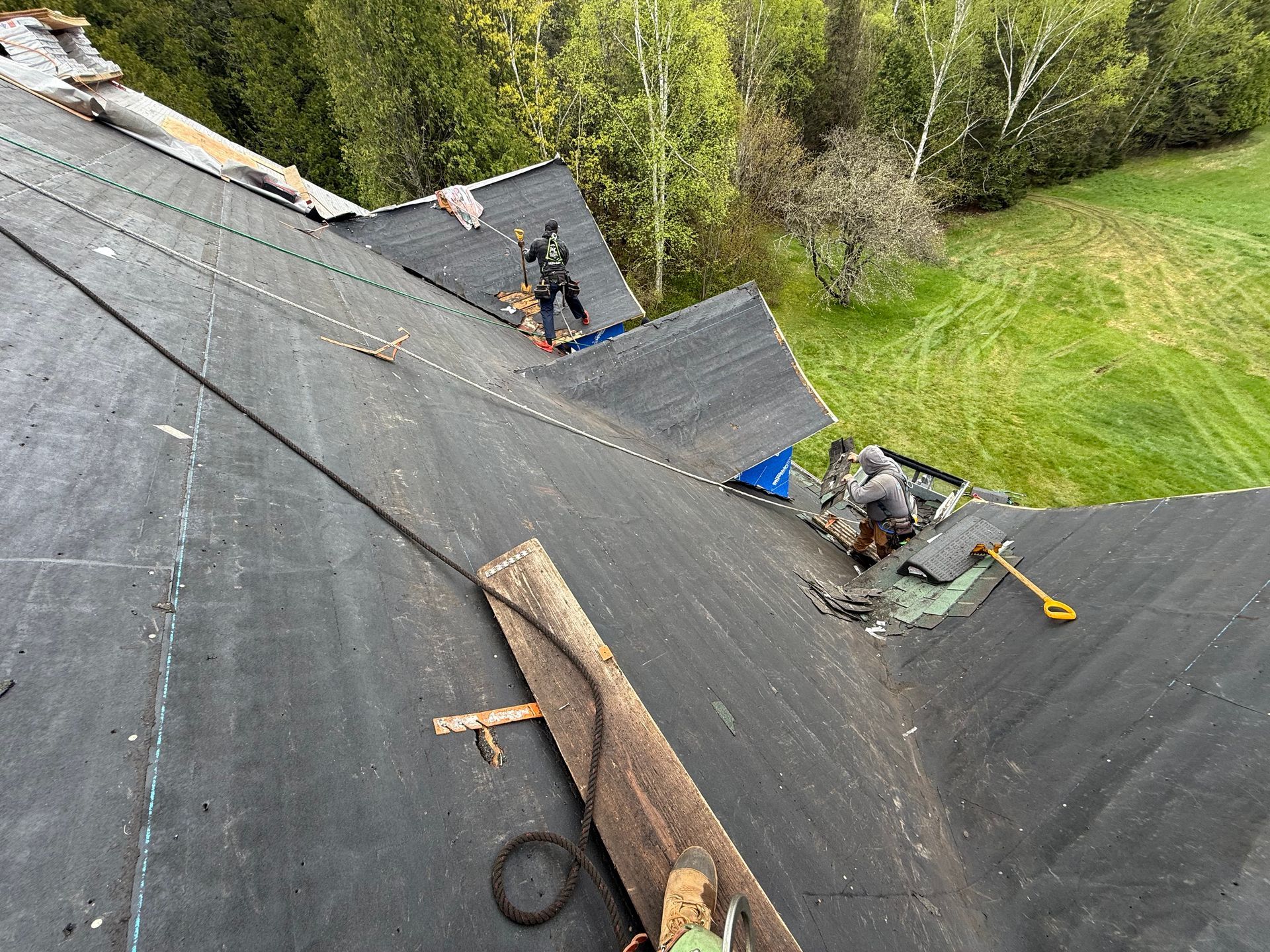Des couvreurs travaillent sur un toit en bardeaux noirs, avec de l'herbe verte et des arbres en arrière-plan.
