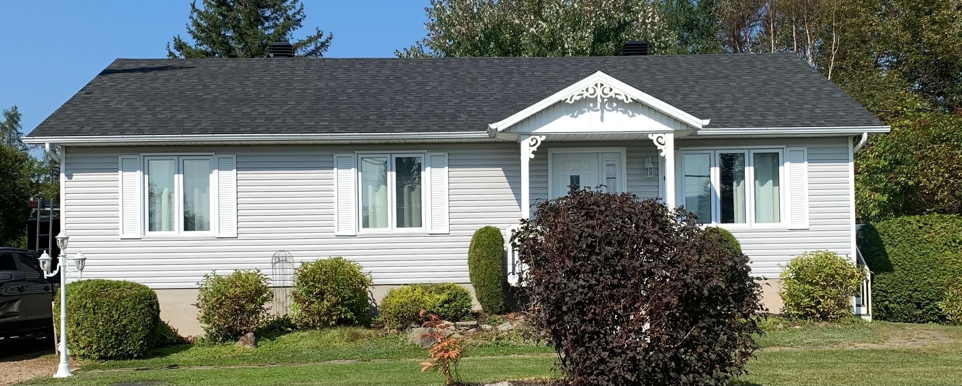 Un bungalow aux façades blanches, au toit foncé, avec des volets et un porche décoratif au-dessus de la porte d'entrée. Aménagement paysager à l'avant.