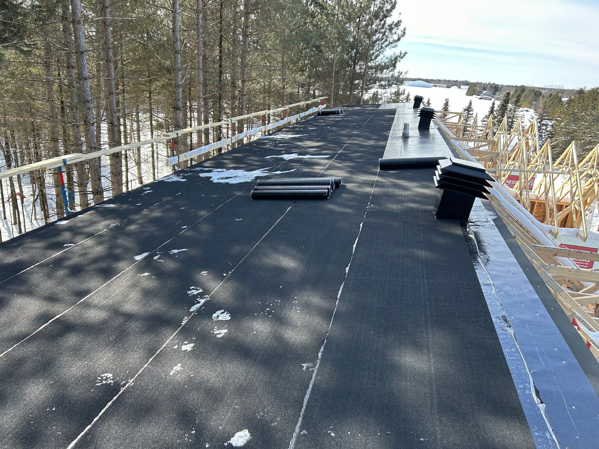 Toit plat recouvert de rouleaux de matériau de couverture noir et de plaques de neige, entouré d'une balustrade en bois et d'arbres.