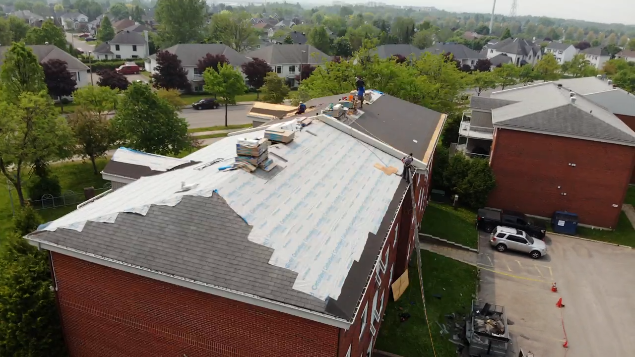 Des ouvriers réparent le toit d'un bâtiment en briques, partiellement recouvert de bardeaux neufs. Vue aérienne, en zone suburbaine.