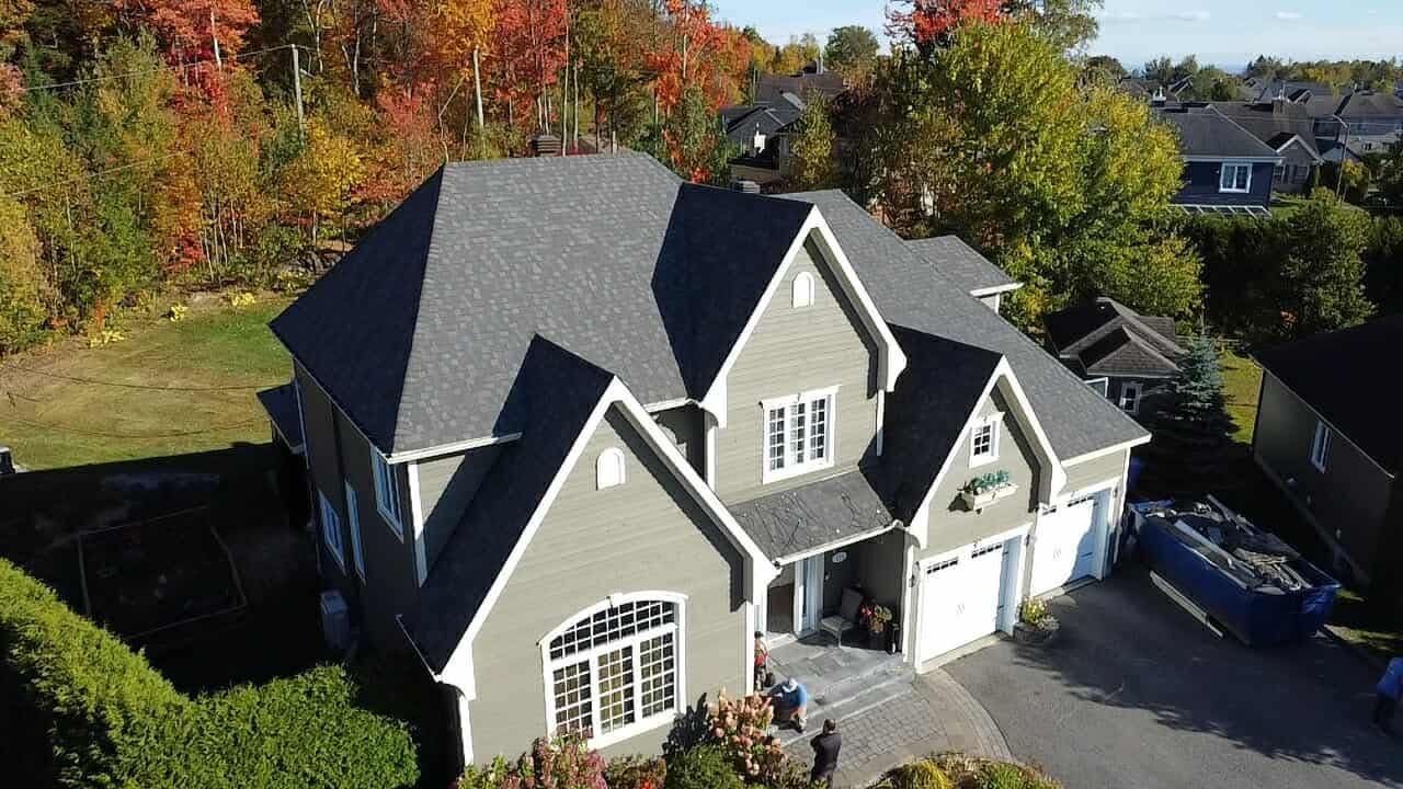 Maison à deux étages avec toit et bardage gris, allée carrossable et feuillage d'automne coloré en arrière-plan.