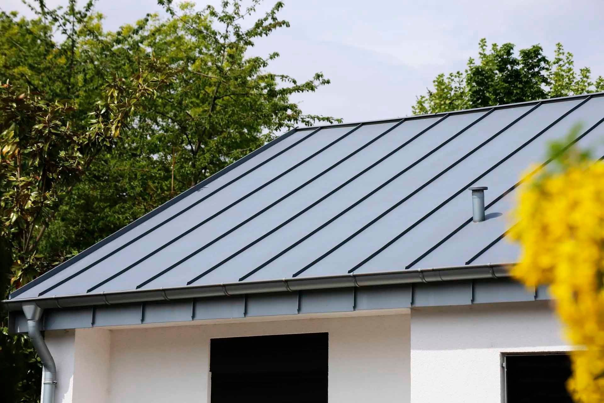 Toit en métal gris sur une maison blanche avec une gouttière et des arbres en arrière-plan.