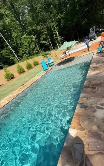 Long narrow blue pool with stone deck and lounge chairs in a leafy backyard