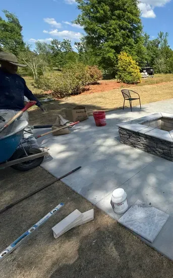 Concrete patio with construction tools, red bucket, chair, and a person working at the left edge outdoors