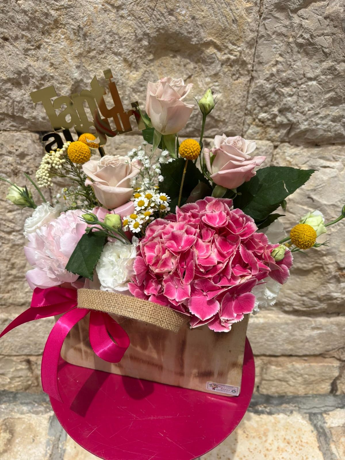 Composizione floreale in un contenitore di carta, nastro rosa, fiori rosa e beige, contro un muro di pietra.