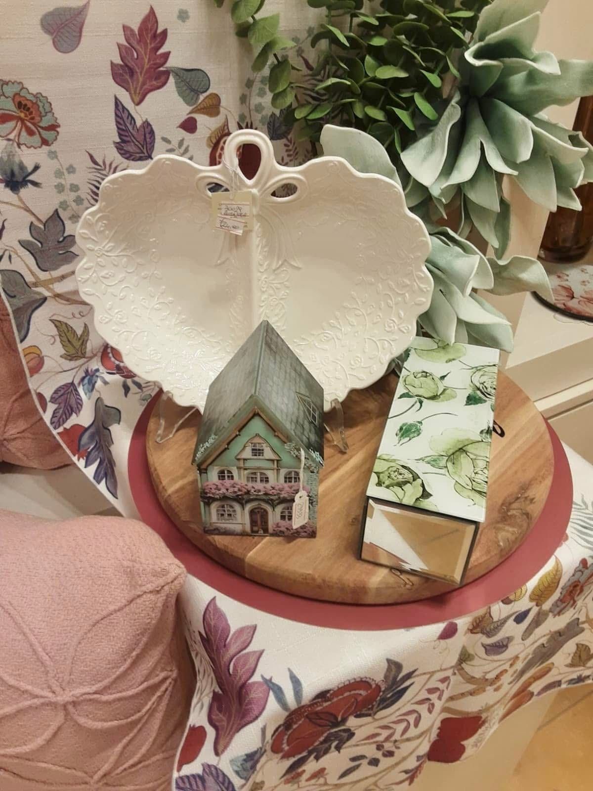 Composizione decorativa su una tavola di legno: un piatto a più livelli, una candela a forma di casa e un regalo incartato, il tutto adagiato su un tessuto floreale.