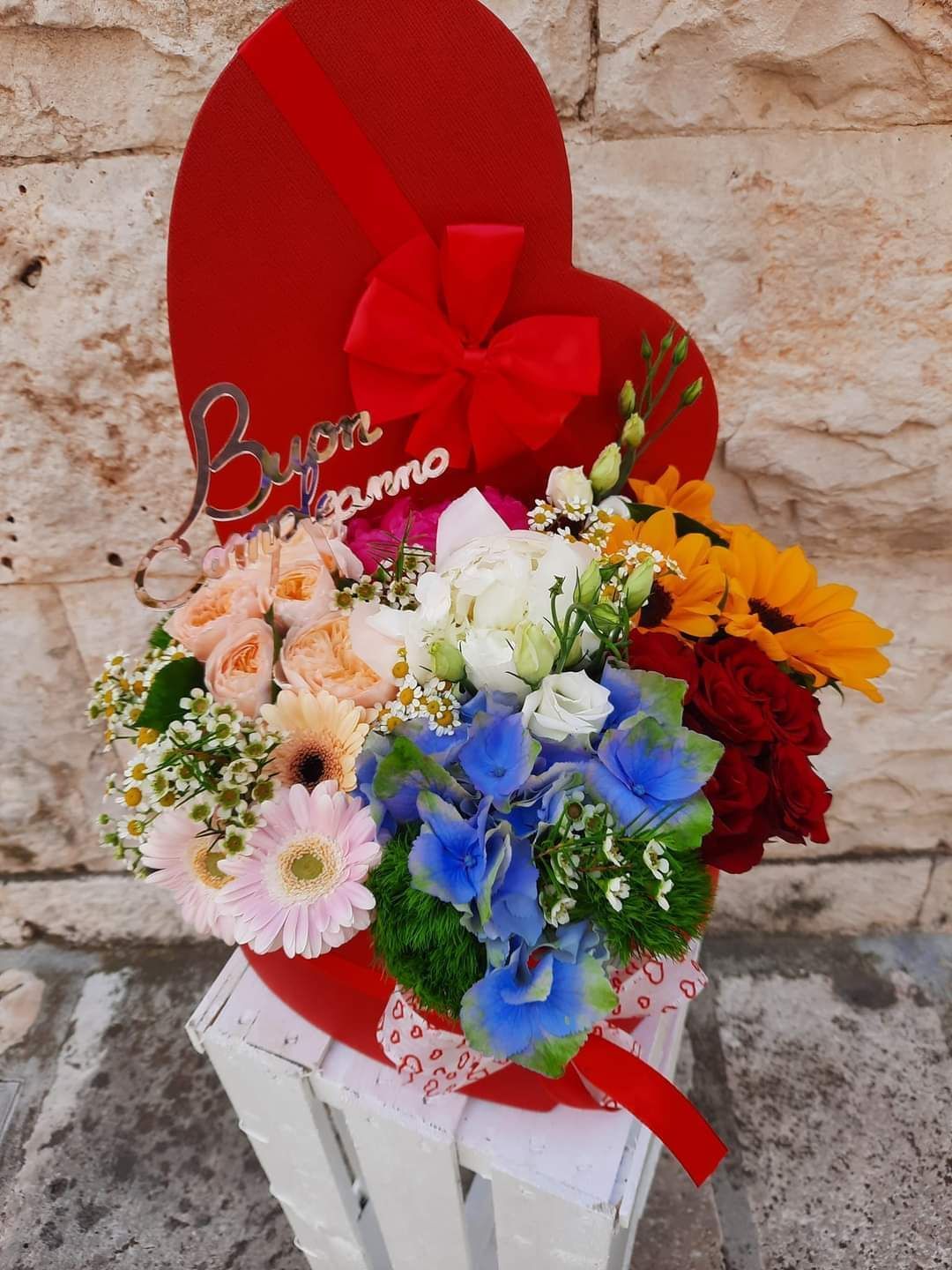 Scatola rossa a forma di cuore con una composizione floreale di fiori colorati e un fiocco di nastro, su una cassa bianca.