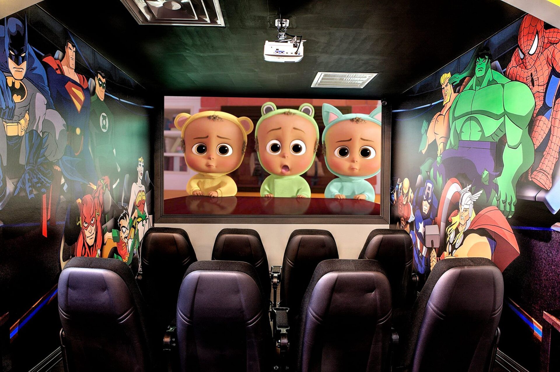 Home theater with rows of seats and colorful superhero wall art, facing a screen showing three cartoon babies