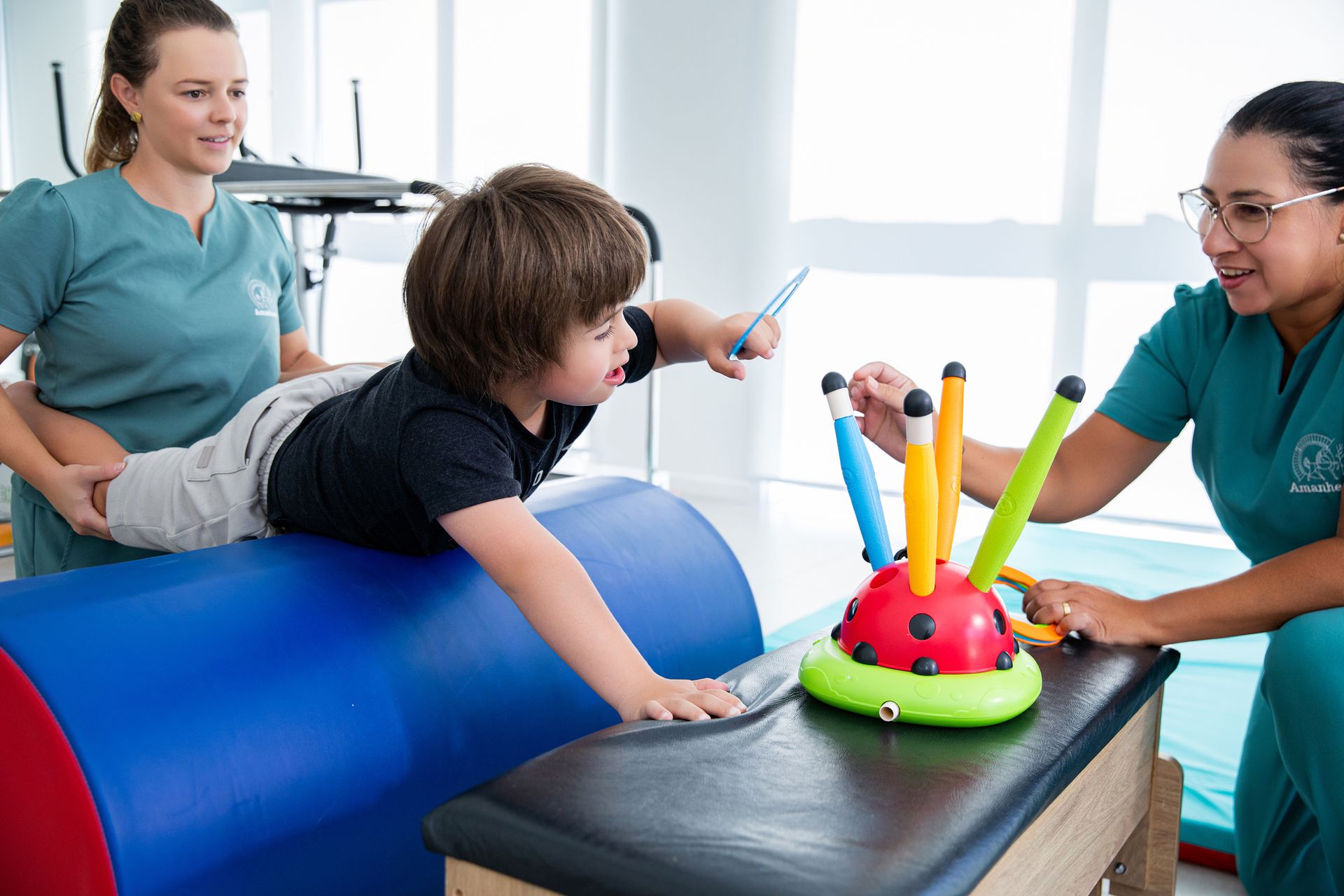 Criança fazendo terapia em um barril de exercícios azul com dois terapeutas em uma sala de clínica bem iluminada.