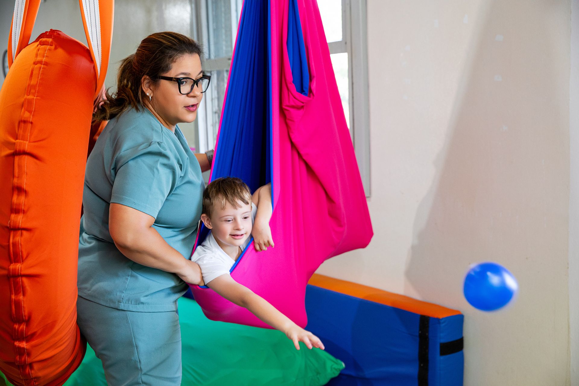 Criança e adulto em balanços terapêuticos coloridos, com uma bola azul por perto, em uma sala de jogos.