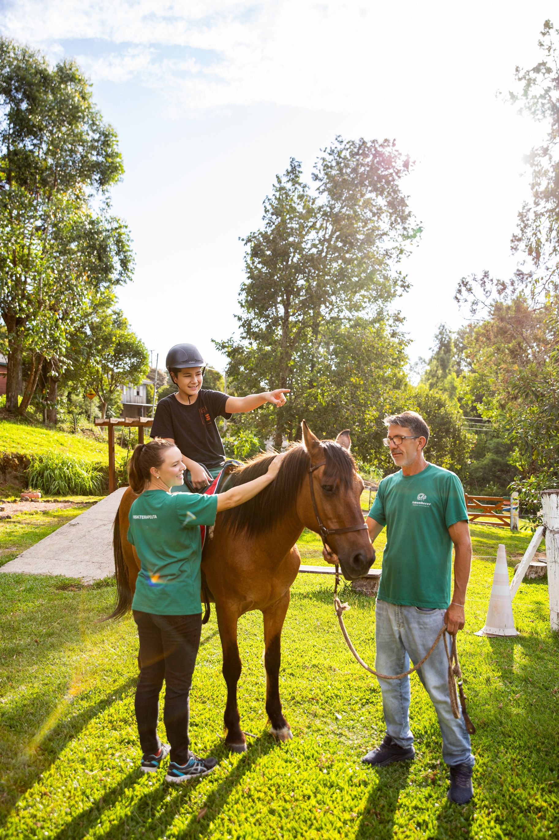 Pessoas acariciando um cavalo marrom ao ar livre em uma trilha ensolarada, com uma pessoa sentada em suas costas.