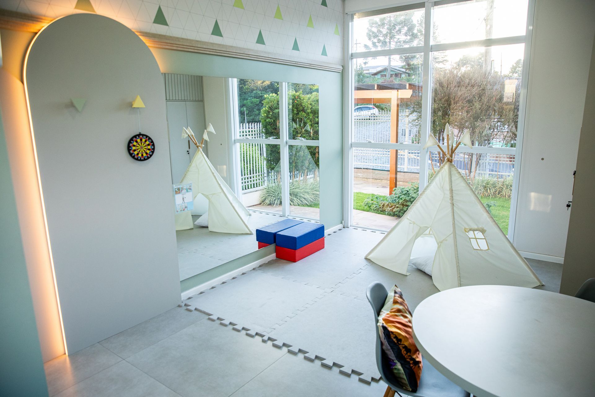 Sala de jogos iluminada com duas tendas brancas, uma parede com alvo de dardos e grandes janelas com vista para um jardim.