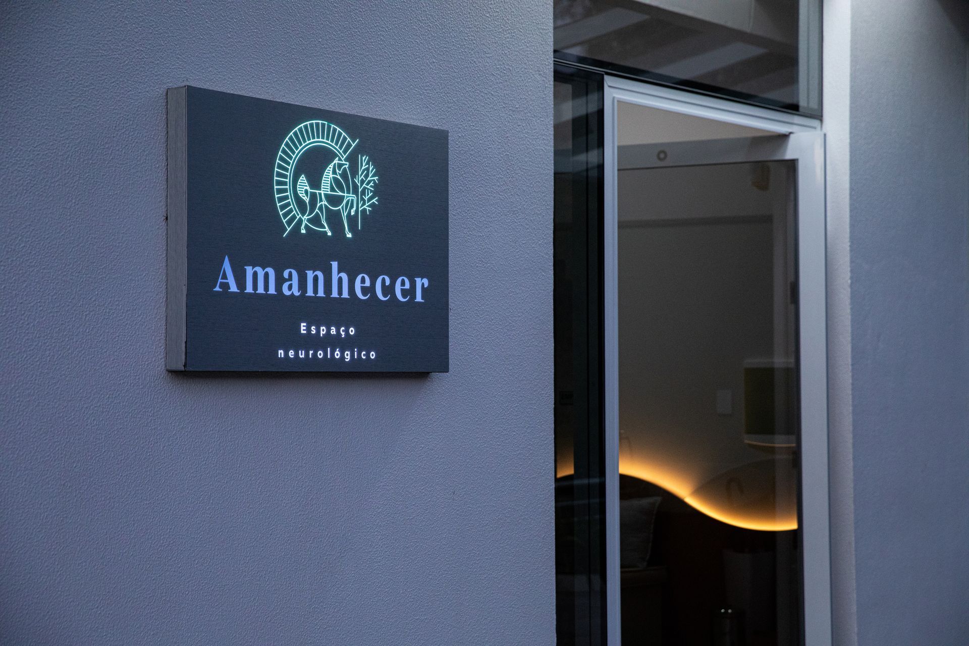 Placa do lounge Amanhecer ao lado de uma porta aberta ao entardecer, com iluminação interna aconchegante visível.