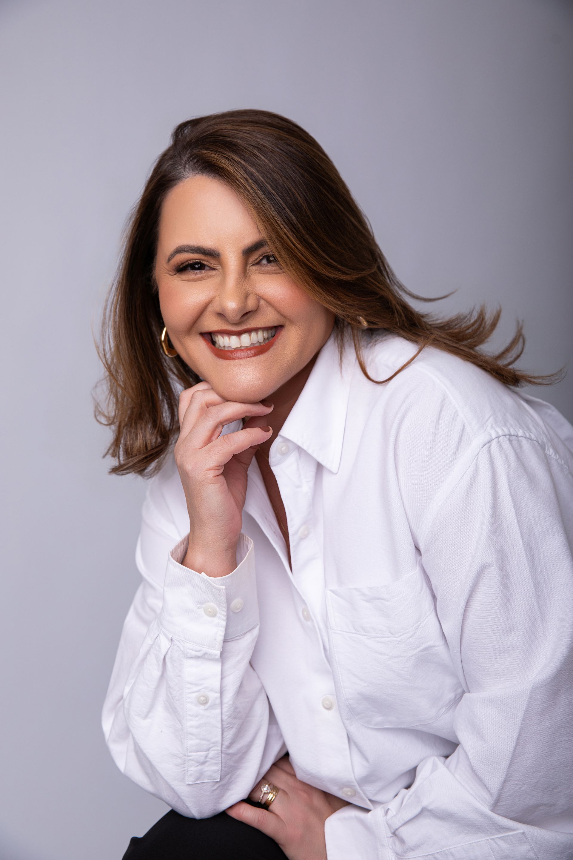 Mulher sorridente de camisa branca apoiada na mão contra um fundo cinza