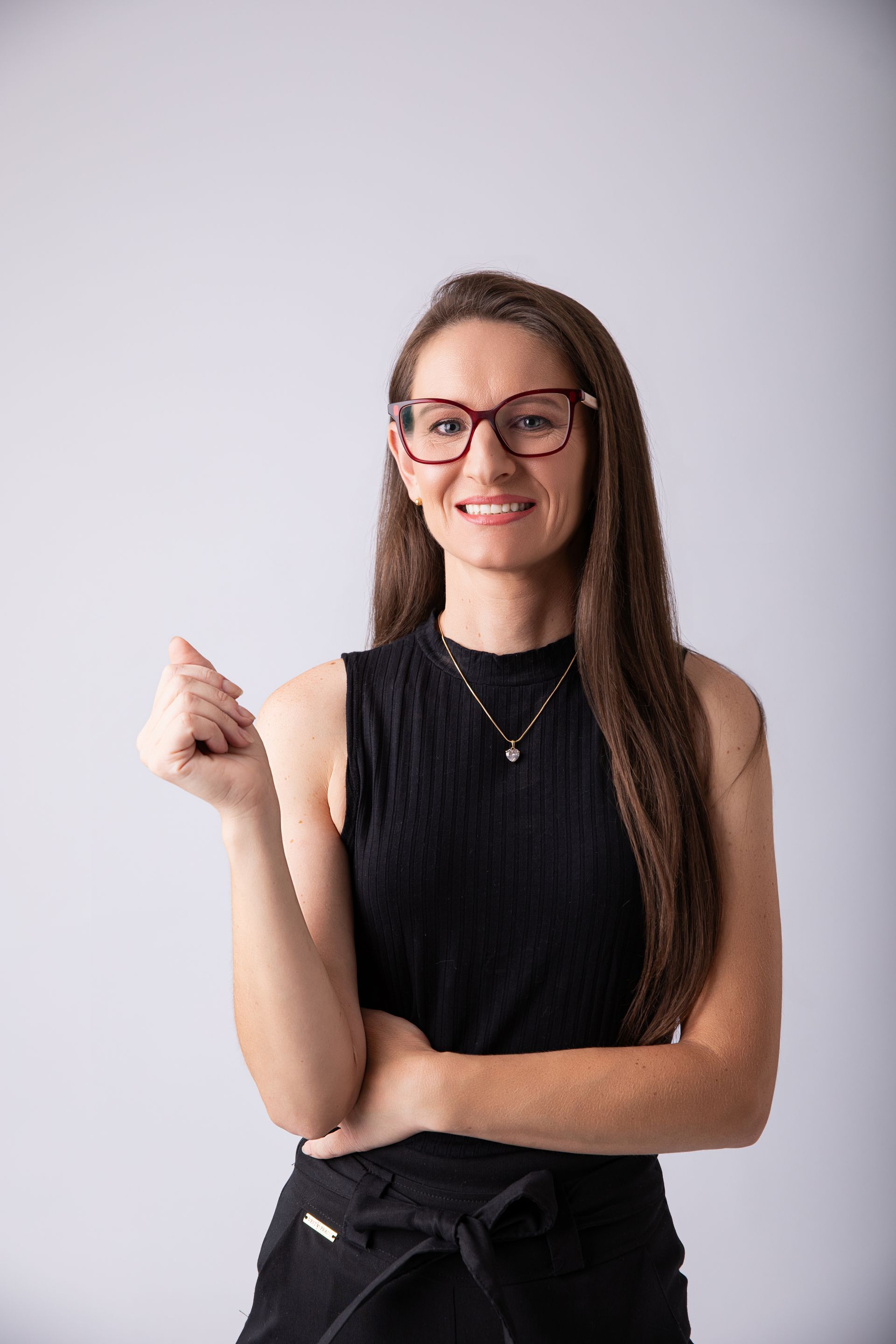 Mulher sorridente de óculos, em pé contra um fundo cinza claro, vestindo uma blusa preta sem mangas.