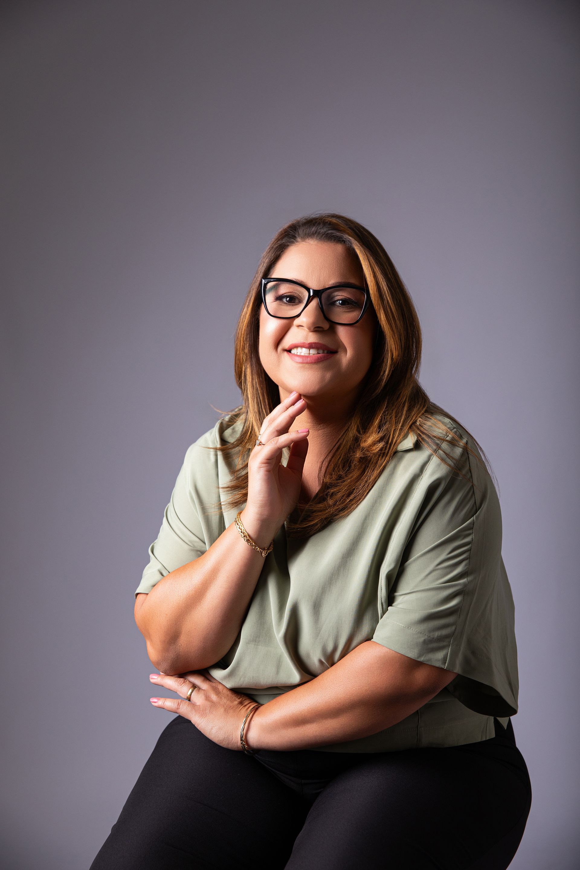 Mulher sorridente de óculos e blusa verde-oliva sentada contra um fundo cinza de estúdio.