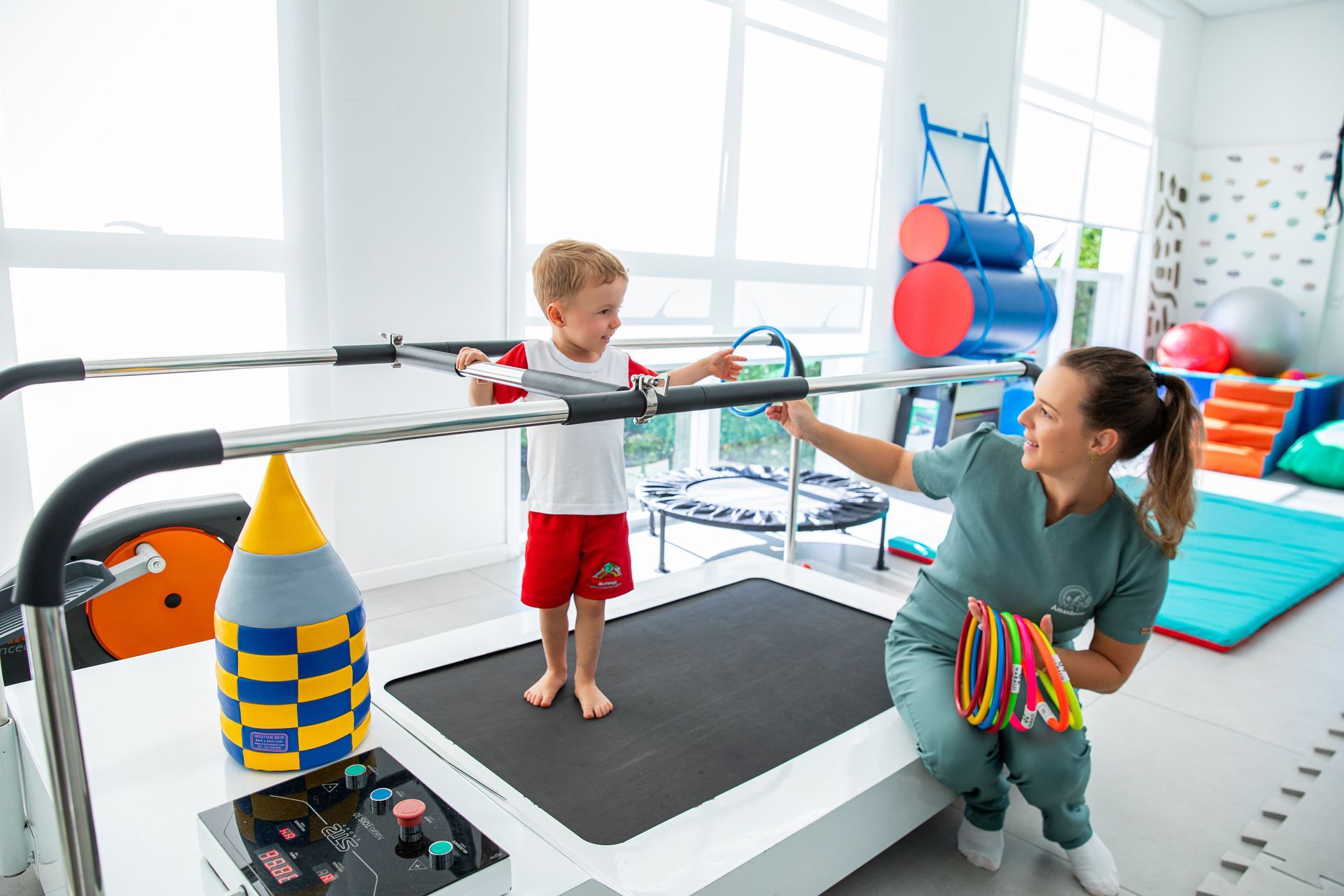 Criança usando barras paralelas com um terapeuta em uma sala de terapia bem iluminada.