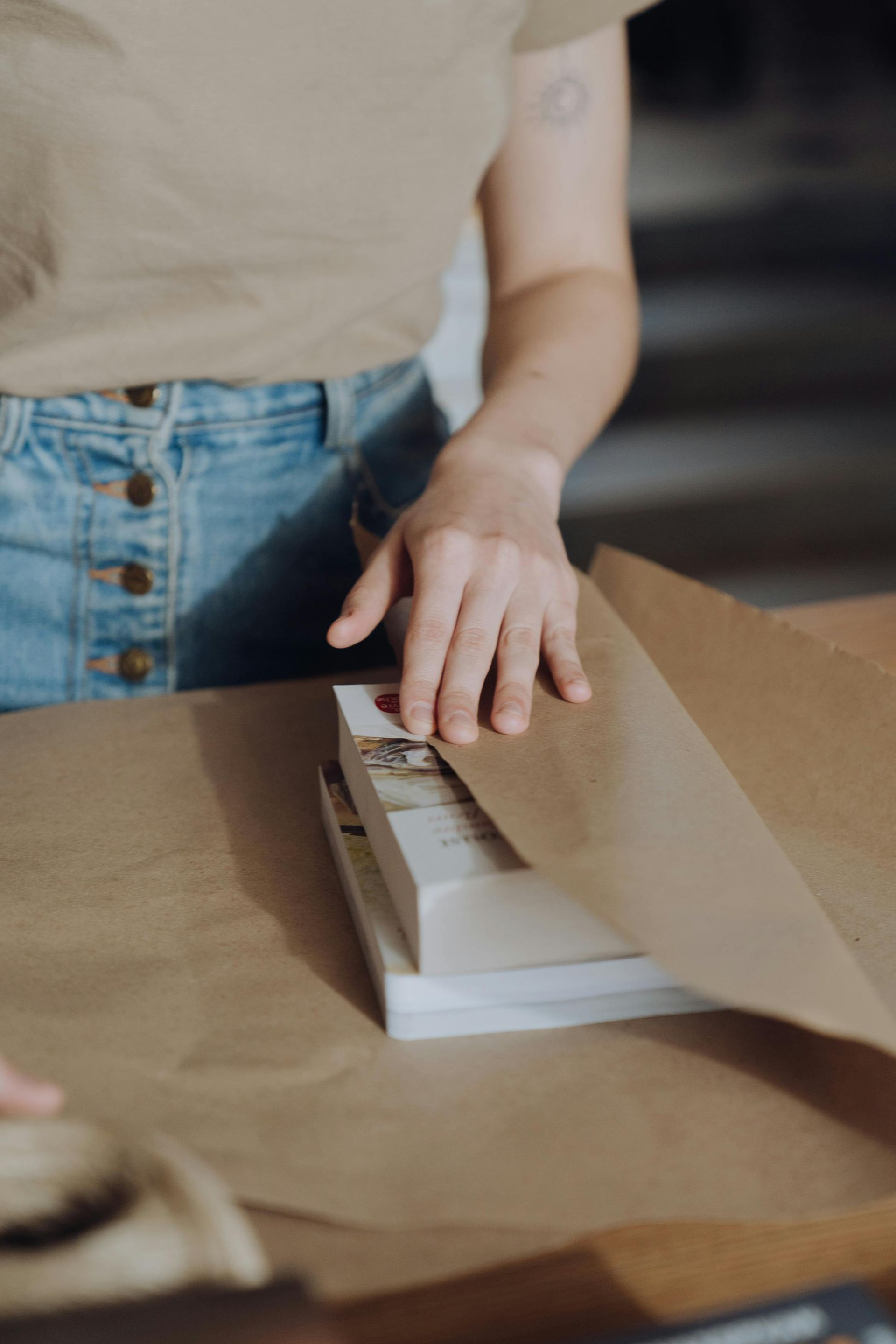 A person wrapping two stacked books in brown paper on a wooden table.