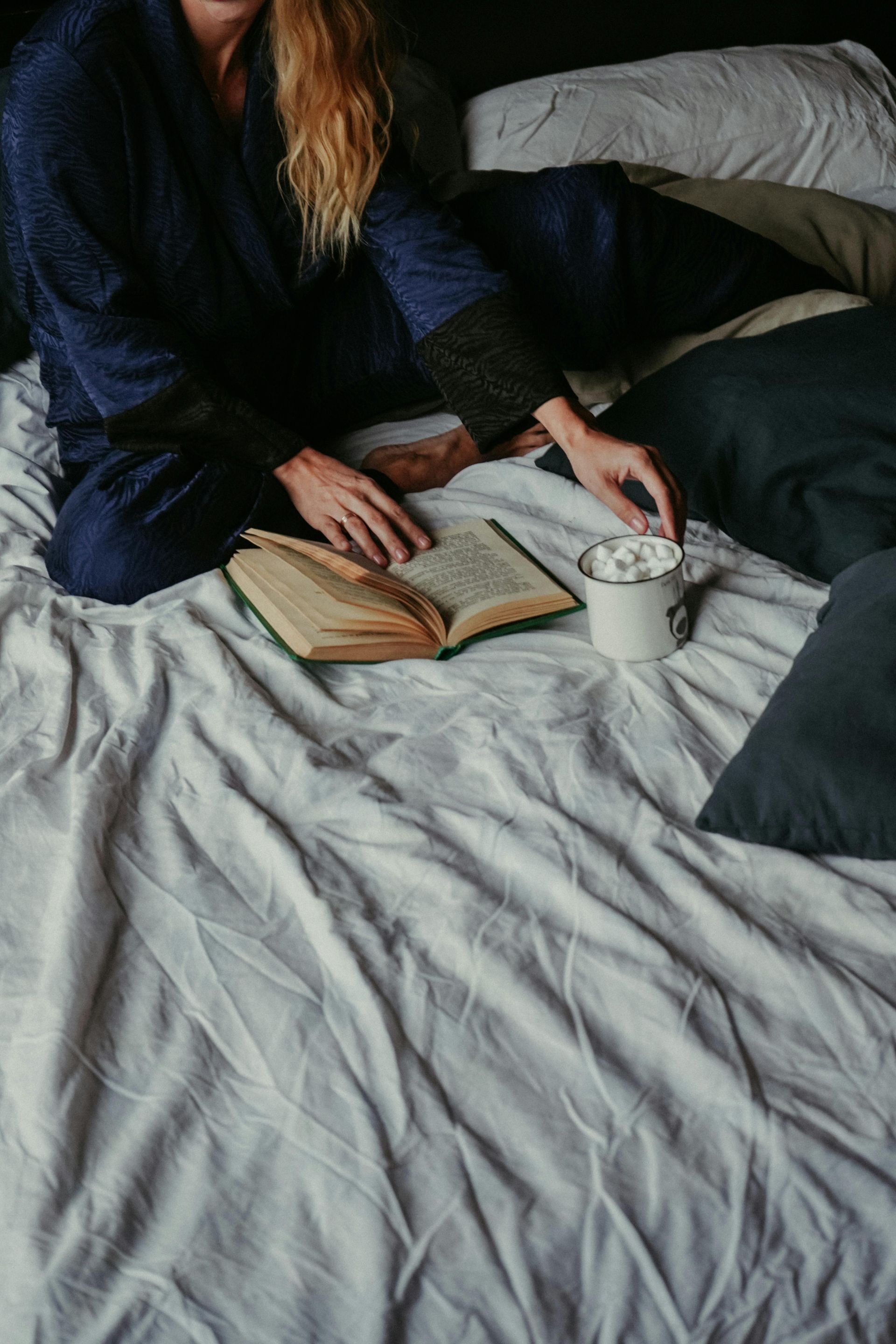 A person in blue loungewear sits on a white bed with an open book and a mug of hot cocoa.