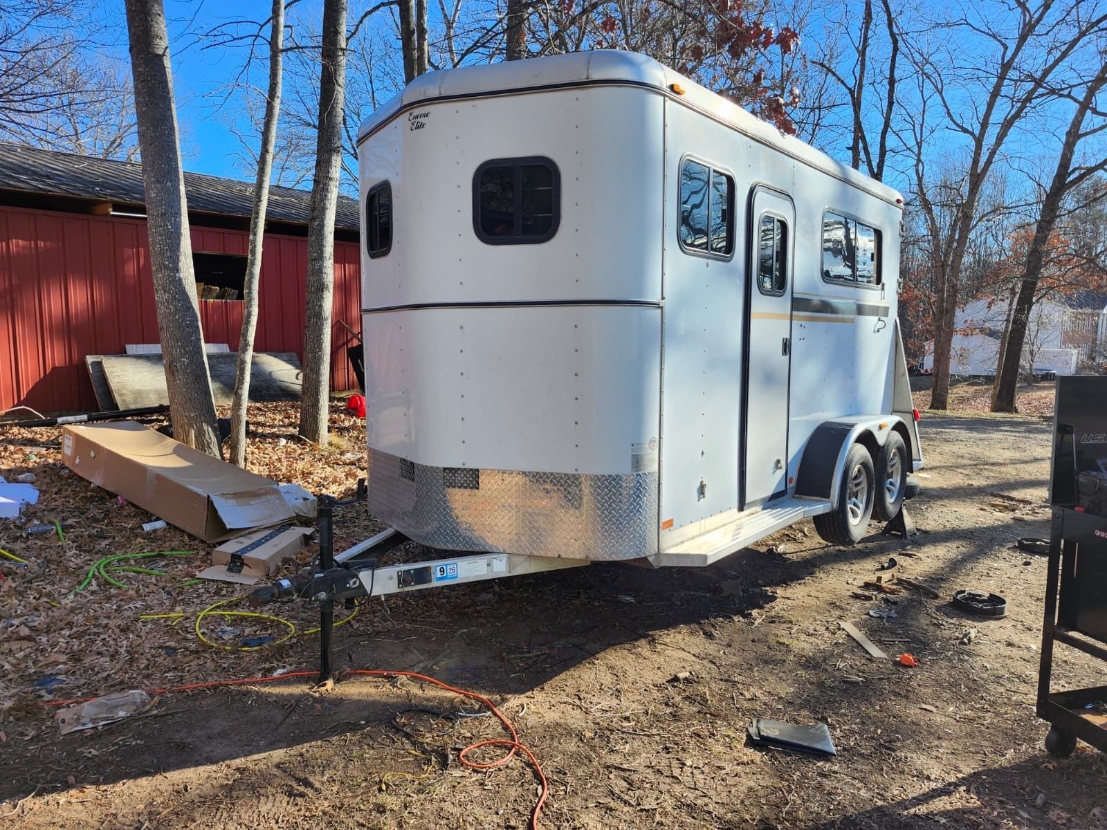 Horse trailer, white with aluminum, parked on a dirt lot near trees and a red building.