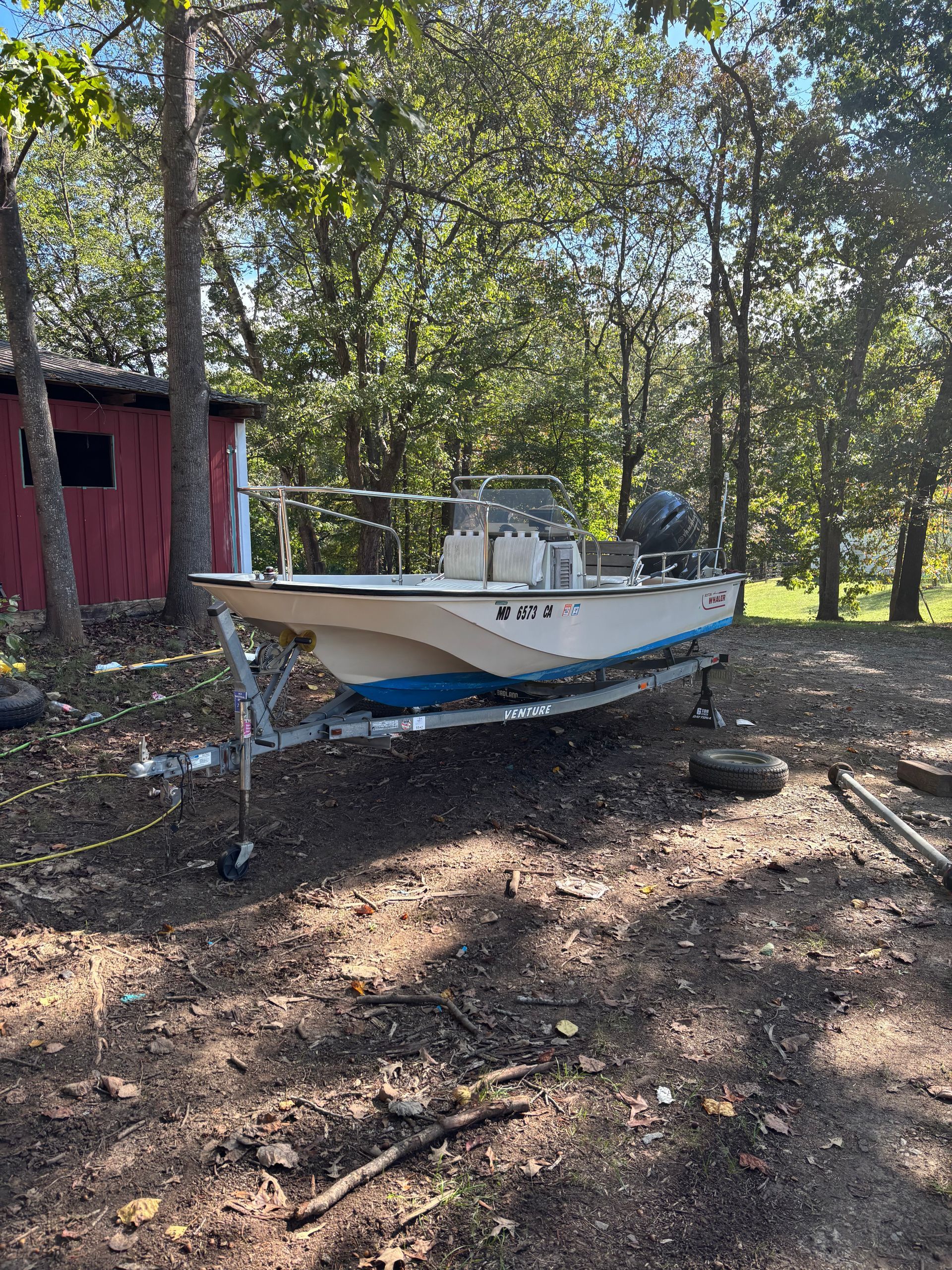 Boat on trailer in wooded area next to a red shed. Boat is white with a blue hull.