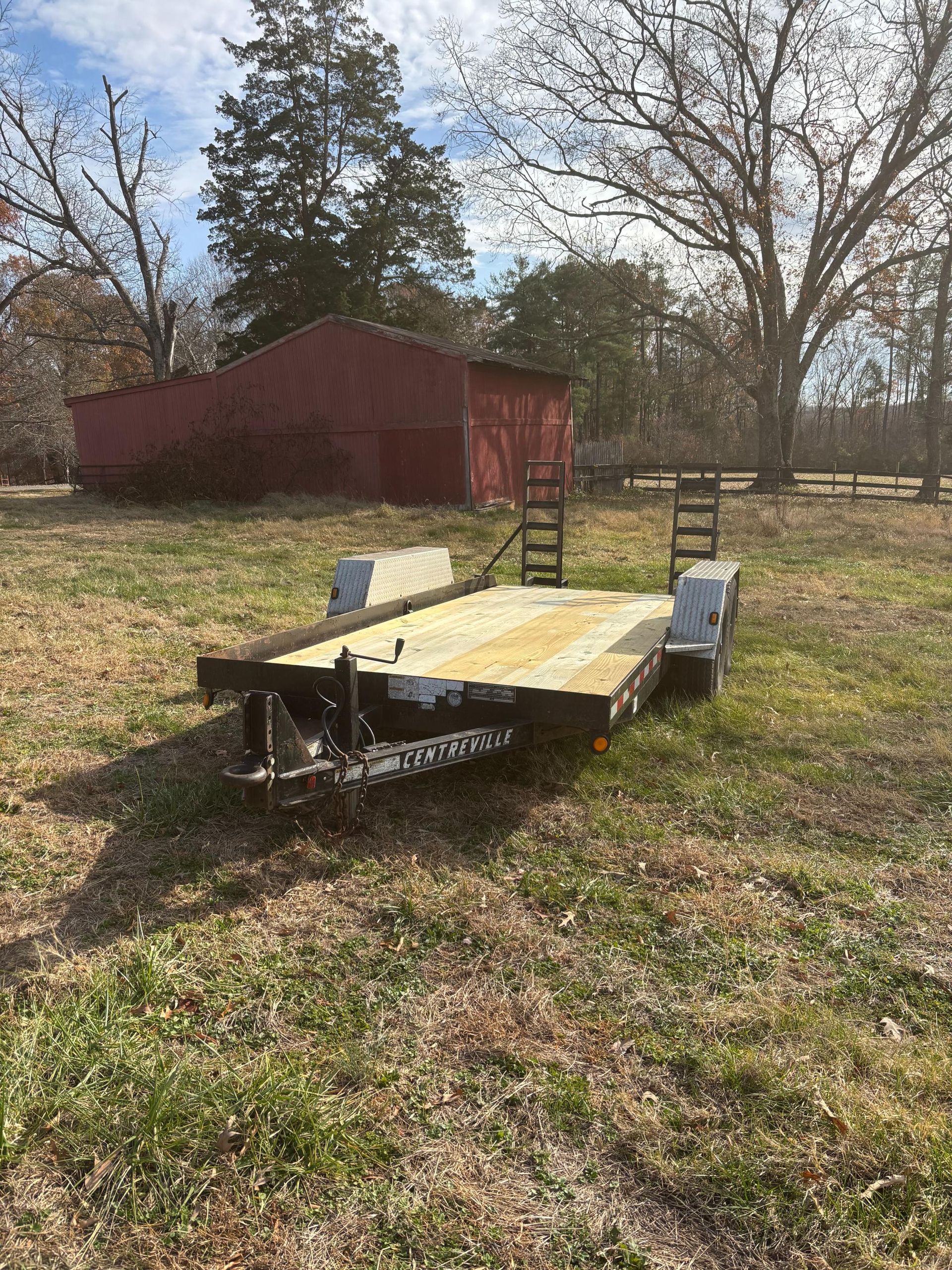 A black trailer with a wooden bed sits in a grassy field in front of a red barn.