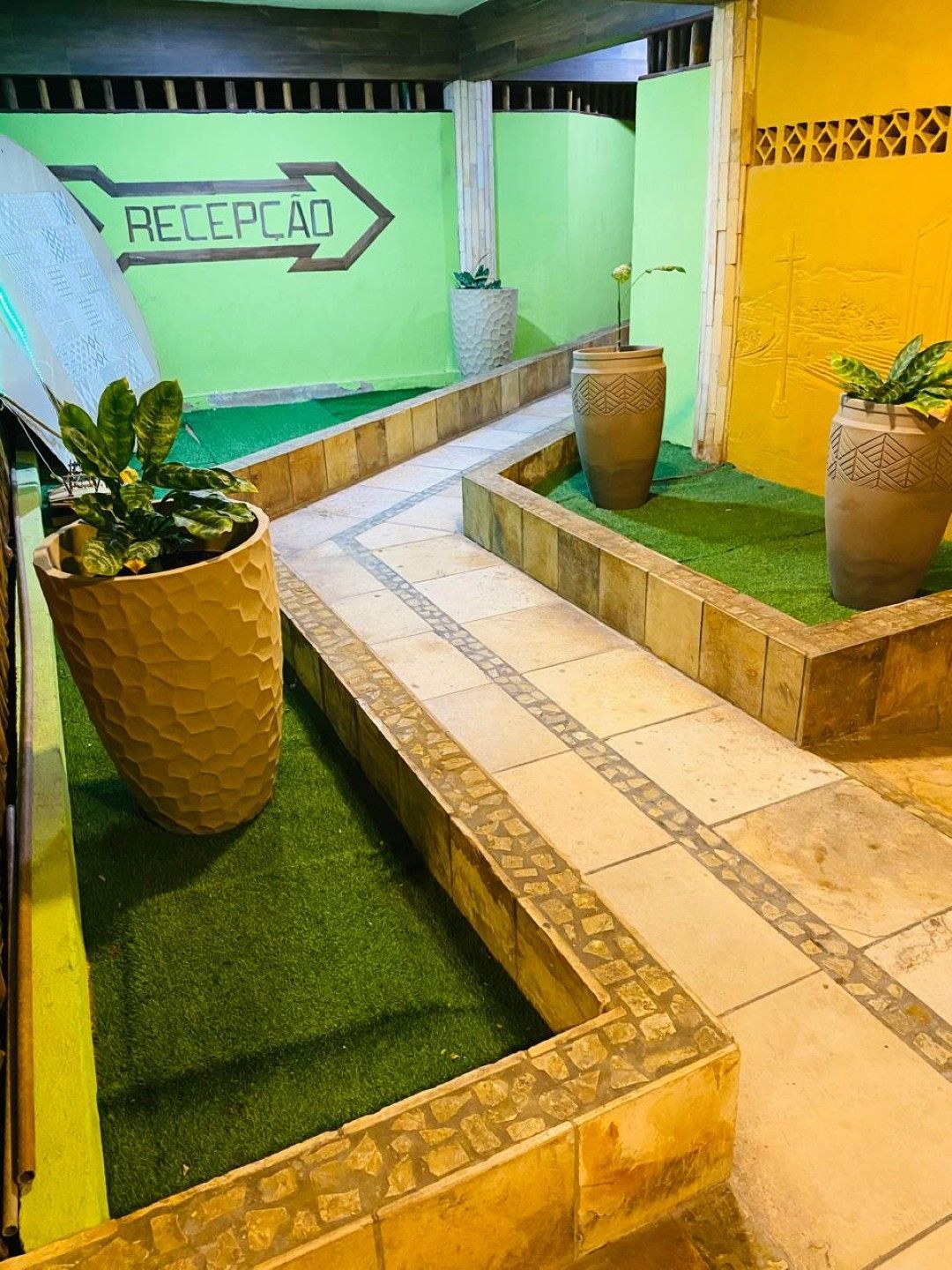 Caminho com piso de azulejos, grama artificial e floreiras. Placa com a inscrição