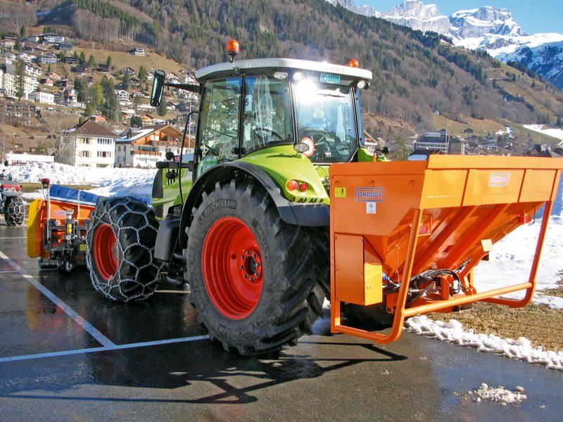 Trattore verde con spazzaneve e spargisale arancioni su una strada di montagna, con cime innevate sullo sfondo.