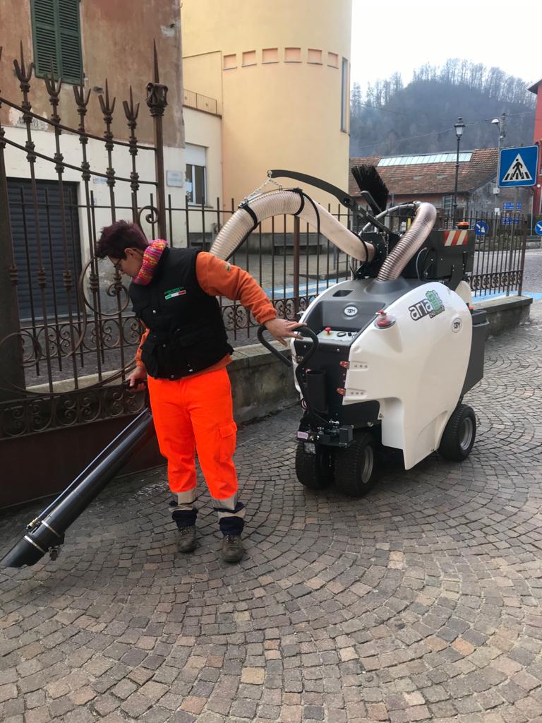 Un addetto alla pulizia stradale utilizza una spazzatrice aspirante bianca su una strada acciottolata accanto a un edificio e a una recinzione.