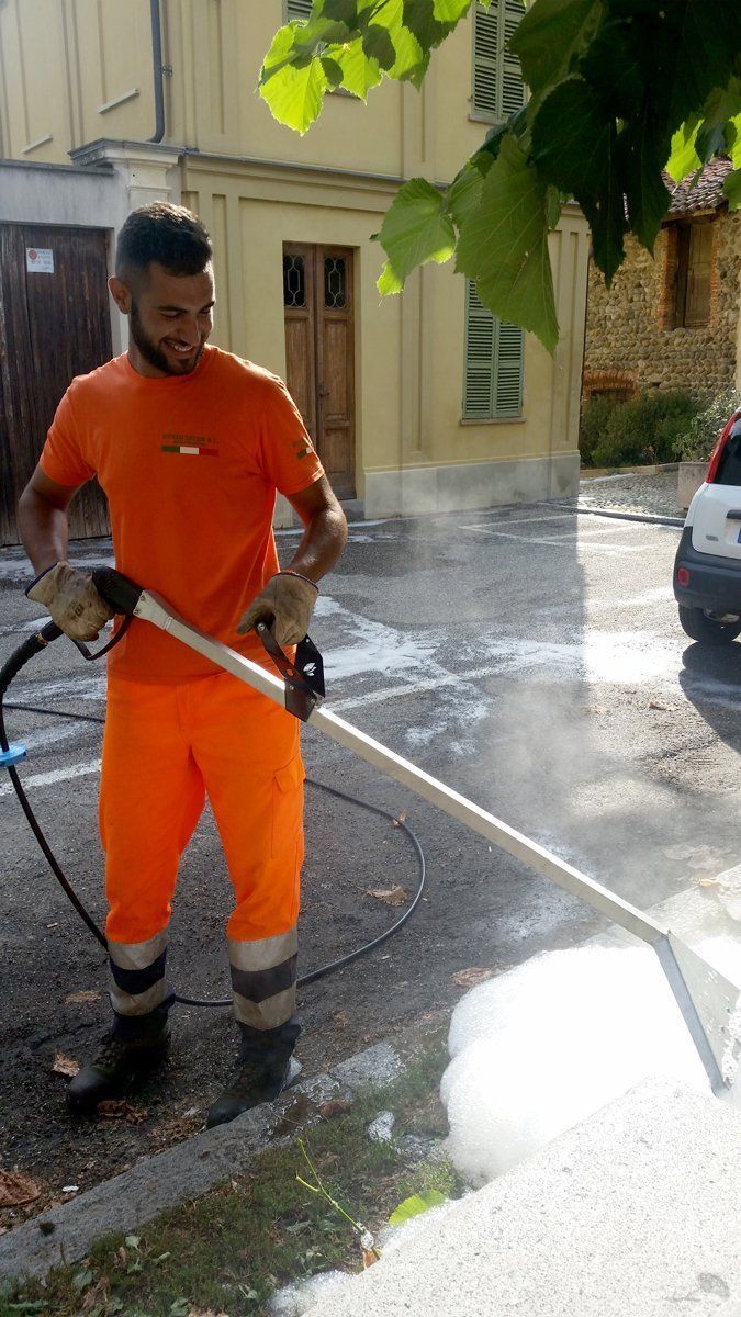 Un operaio in uniforme arancione pulisce un marciapiede con l'idropulitrice in una strada soleggiata.