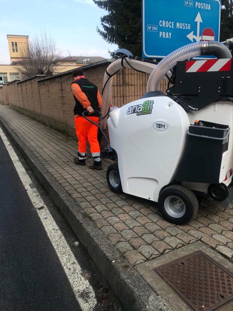 Un operaio con indosso una tuta di sicurezza arancione utilizza una macchina per la pulizia stradale su un marciapiede accanto a un segnale stradale.