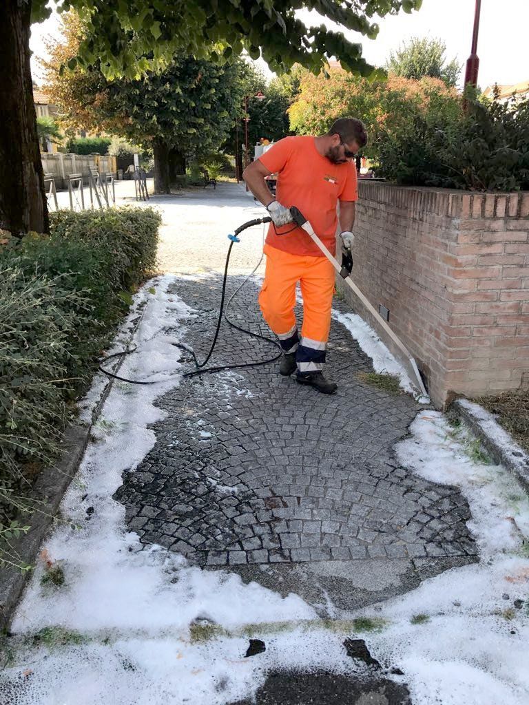 Un operaio con indosso una tuta di sicurezza arancione pulisce con idropulitrice un marciapiede crepato accanto a un muro di mattoni e ad alcuni alberi.
