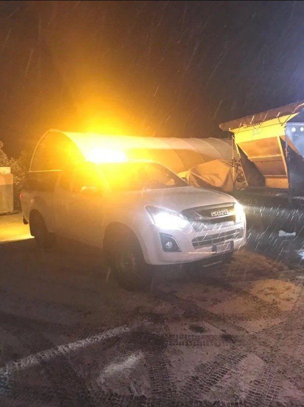 Un pick-up bianco con luci ambra lampeggianti in una notte nevosa accanto a una struttura crollata.