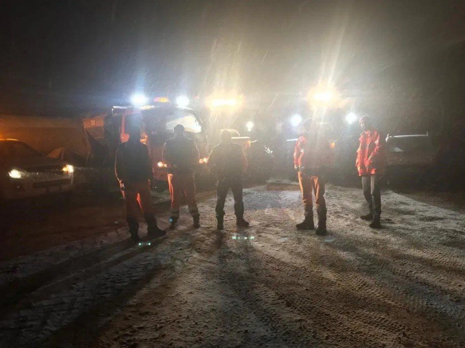 Cinque persone sono in piedi su una strada sterrata di notte, illuminate dai fari abbaglianti e dalle luci dei veicoli.