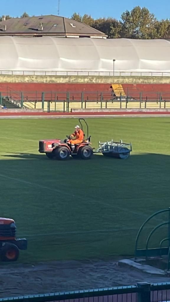 Un operaio falcia un campo sportivo con un tosaerba a motore, con tribune vuote sullo sfondo.
