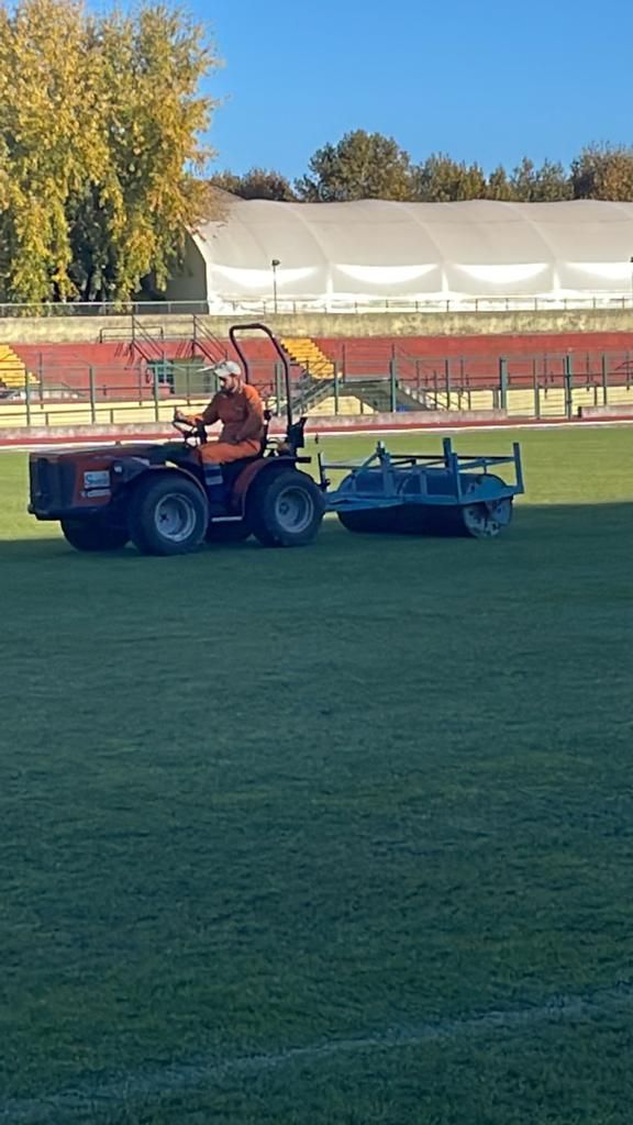 Un trattore arancione falcia un campo verde in uno stadio, con tribune e tendoni sullo sfondo.