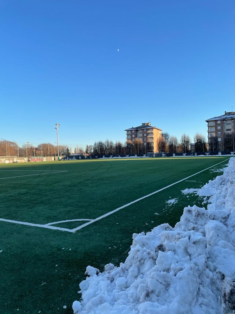 Campo sportivo innevato sotto un cielo azzurro e limpido, con palazzi residenziali sullo sfondo.