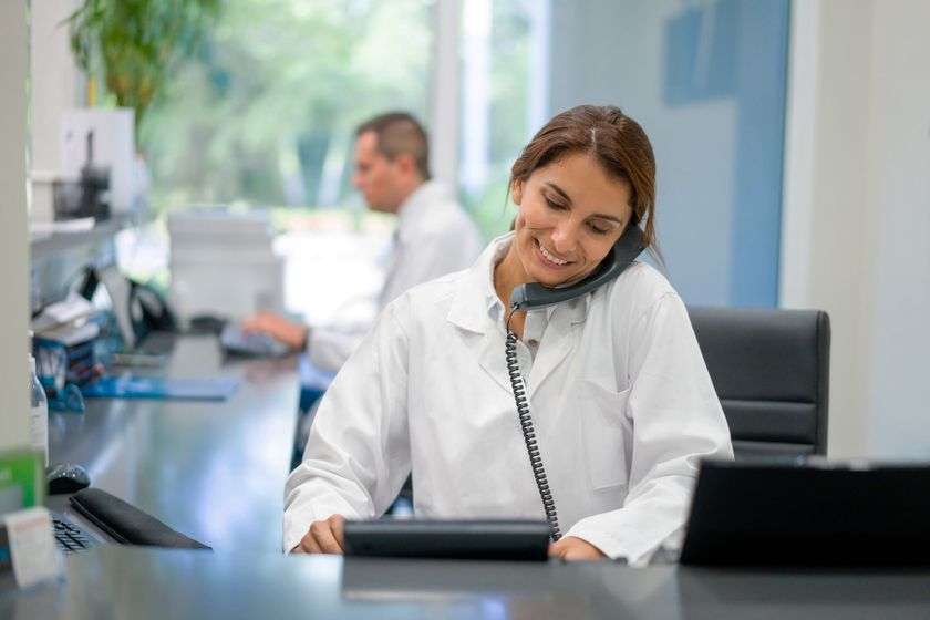 A woman in a lab coat is sitting at a desk talking on a phone.