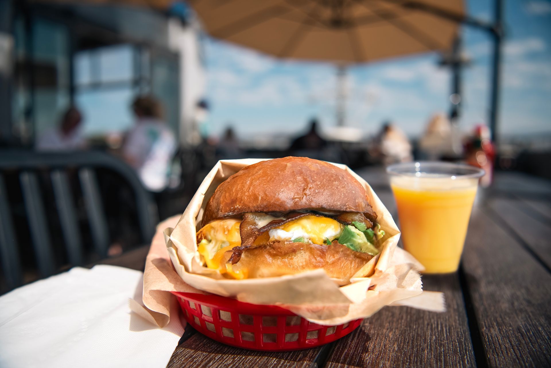 A breakfast sandwich in a red plastic basket next to a glass of orange juice on a wooden table at an outdoor patio.